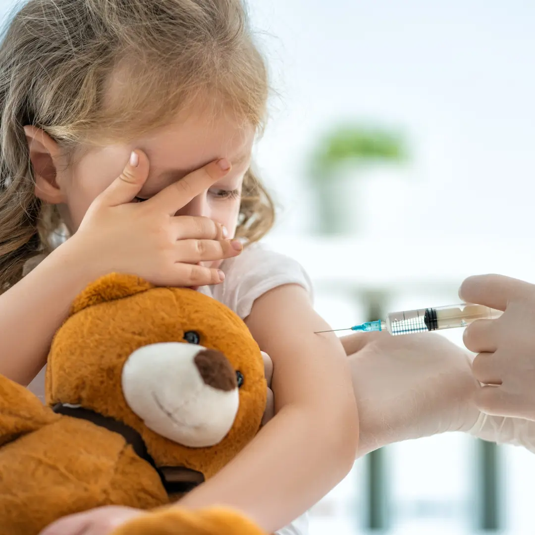 Niña pequeña abrazada a un osito de peluche mirando curiosa mientras le ponen una vacuna. Calendario vacunas pediatria